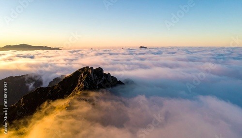 Aerial view of a rocky mountain peak emerging from a sea of clouds under a clear blue sky at sunrise or sunset, with distant mountains visible.