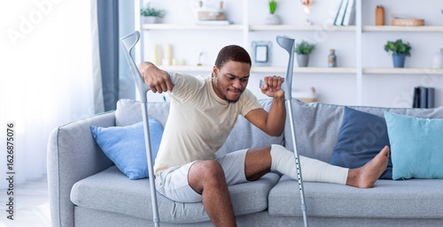 Tablou pe pânză Young black guy with plastered leg leaning on crutches, trying to get up from sofa at home, full length