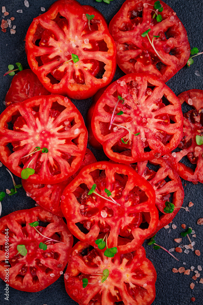 Freshly sliced ripe tomatoes arranged on a textured dark surface with herbs and salt scattered around Freshly sliced ripe tomatoes arranged on a textured dark surface with herbs and salt scattered around