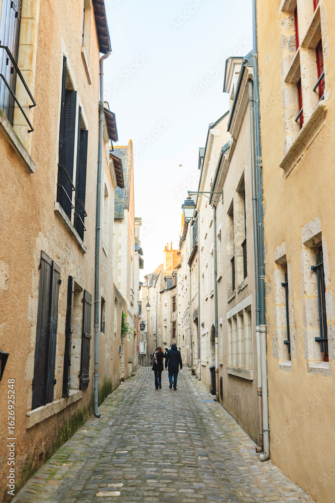 Fototapeta premium Two people stroll down a narrow, cobblestone-paved lane in Tours. Ancient buildings with closed shutters stand on either side. It is an overcast day in early spring