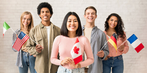Young students of language school celebrate holiday. Happy millennial peoples of different nationalities have fun on study and hold little flags on brick white wall background, studio shot, free space
