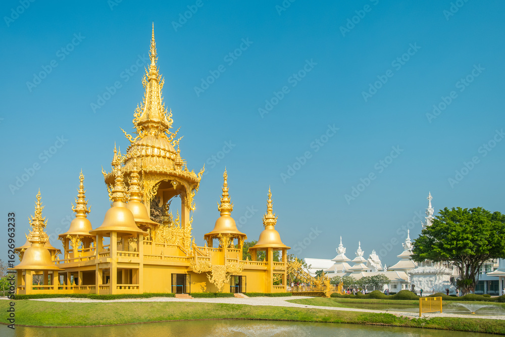 Naklejka premium Golden Ganesha temple in Wat Rong Khun complex with ornate spires and traditional architecture surrounded by garden and pond under blue sky in sunny day, Chiang Rai, Thailand. White temple complex