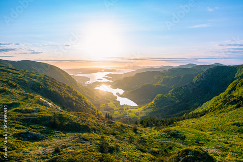 Lush green tree, grass and marsh lined valley leading into the fjord surrounding the nordic city Bergen, Norway, at the start of a midnight sunset with an overpoweringly strong sun