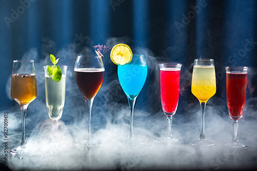 A row of colorful cocktails in elegant glasses with smoke or mist around them, on a reflective surface with a dark blue curtain background