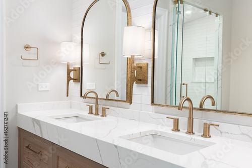A bathroom detail with gold faucets, light fixtures, and mirrors, a wood cabinet, large marble slab countertop, and a subway tile shower.