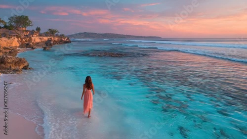 A woman in a flowing dress walks on a pink beach as the sun sets over the ocean, cliffs in background