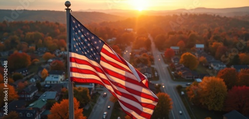Fototapeta Naklejka Na Ścianę i Meble -  Aerial view of small American town with US flag flying. Autumn foliage surrounds rolling hills, houses, streets under serene sunset sky. Peaceful rural community lifestyle with patriotic pride,