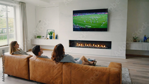Cozy Moments in a Living Room: Three individuals unwind in a modern living room, eyes fixed on the large television screen above a contemporary fireplace, enjoying their leisure. 