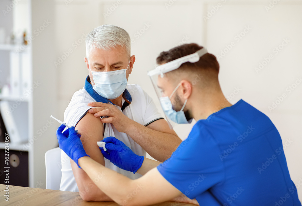 Fototapeta premium Young doctor making coronavirus vaccine injection to senior male patient at hospital. Mature man being immunized against covid-19 at clinic. Medical treatment against global viral epidemic concept