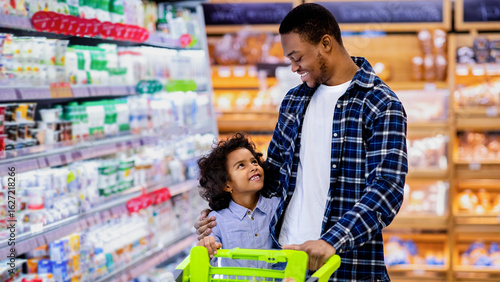 Tableau sur toile Happy black dad and his pretty little daughter with shopping trolley having great time buying groceries at big mall