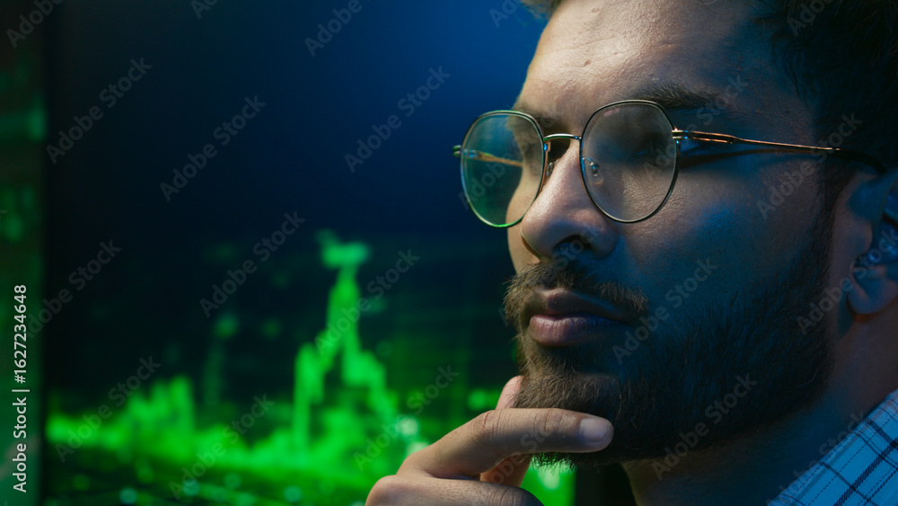 Fototapeta premium Indian Arabian man in glasses thinking contemplate ponder solving coder trader IT specialist male ethnic guy businessman business programmer analysing trading chart graph crypto currency neon indoor