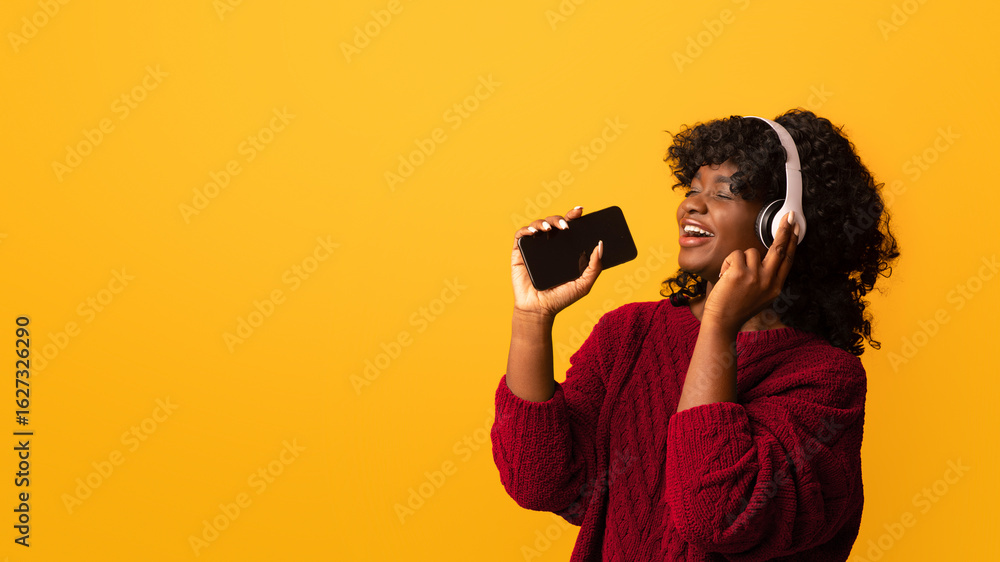 Joyful curly african american woman with smartphone and wireless headset singing on yellow studio background, using mobile phone as microphone, panorama with copy space. Entertainment concept Joyful curly african american woman with smartphone and wireless headset singing on yellow studio background, using mobile phone as microphone, panorama with copy space. Entertainment concept