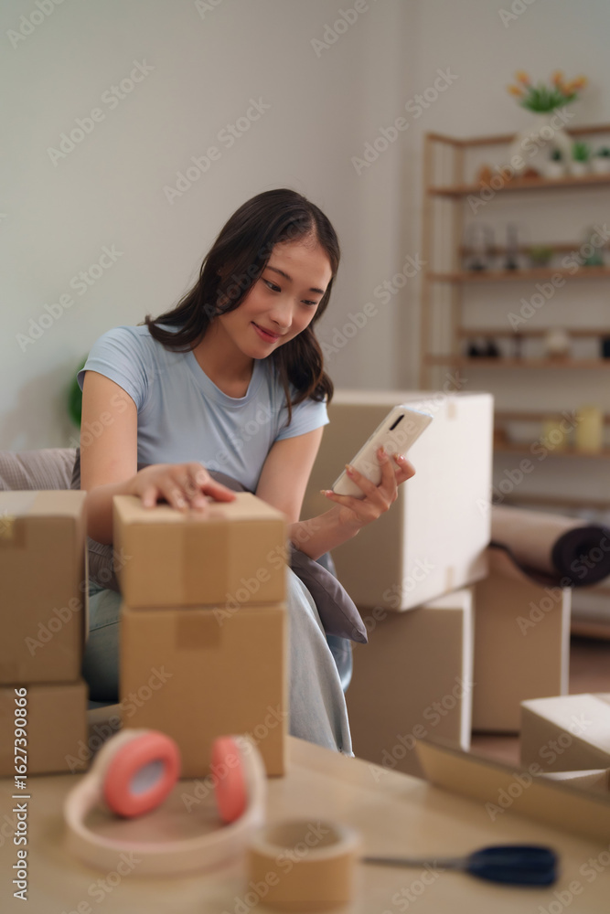 Asian young adult woman packing moving boxes at home checking smartphone organizing belongings for relocation or managing small business operations Asian young adult woman packing moving boxes at home checking smartphone organizing belongings for relocation or managing small business operations
