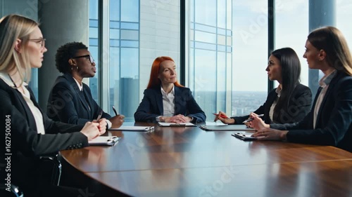 video of a female-led executive boardroom meeting bathed in natural light. Five confident women in sharp business attire sit around a polished mahogany table, engaged in serious discussion
