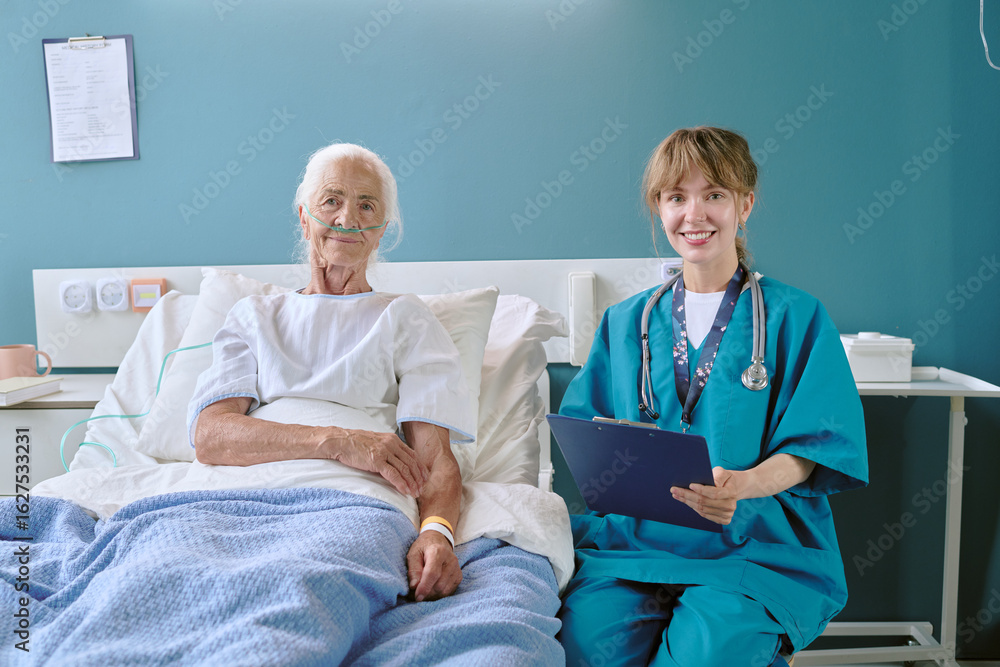 Obraz premium Senior Caucasian woman lying in hospital bed with oxygen tube, smiling young adult Caucasian female nurse sitting beside bed holding clipboard, both looking at camera