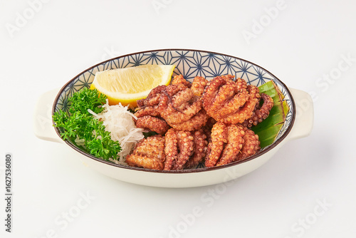 Crispy fried octopus tentacles served in decorative bowl with fresh parsley, shredded radish, lemon wedge, and banana leaf garnish, offering vibrant and appetizing presentation