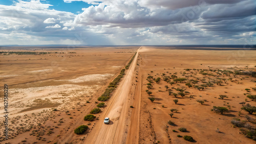 The road is flanked by sparse vegetation and stretches towards the horizon under a partly cloudy sky. A single vehicle, leaving a trail of dust, travels along the road.