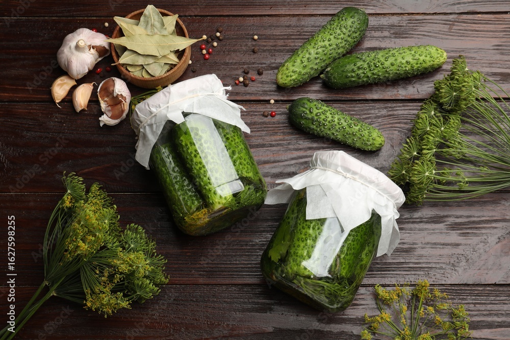 Fototapeta premium Pickled cucumbers in jars, fresh ones, garlic, dill and dry bay leaves on wooden table, flat lay