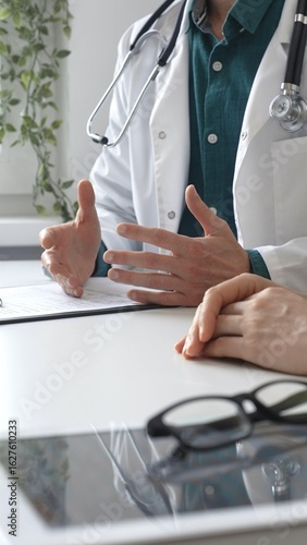 Doctor man wearing medical coat over green shirt, and stethoscope is gesturing with hands while explaining diagnosis to patient during medical consultation in clinic office. Medicine and health care