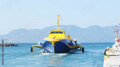 Wallpaper Mural High-speed hydrofoil catamaran in yellow and blue colors leaving the port of Aegina, Greece. Crew member on board. Visible mooring elements, lighthouse, mountains and clear sky in background. Torontodigital.ca