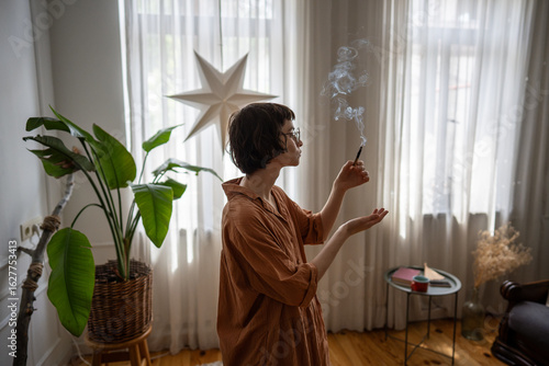 Focused young woman do personal ceremony with smudging stick to refresh and enhance mental clarity. Aromatherapy benefit, spiritual connection, home sanctuary creation, slow living, sensory therapy