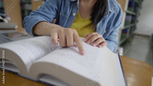 A focused young Asian female student with glasses uses a laptop and books to do research in a library, representing digital learning, hybrid education, and student lifestyle concepts.