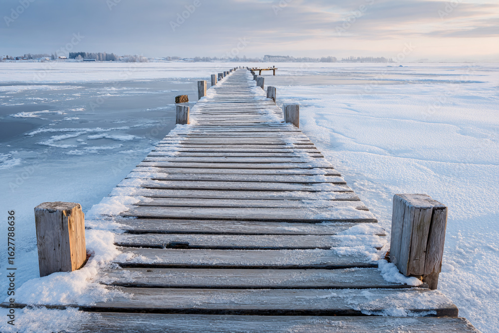 Obraz premium Wooden pier covered in snow leading to frozen lake