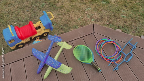 Backyard photo with a pile of outdoor toys for kids. A bunch of sports toys are lying on a wooden patio. Tennis rackets, plastic hoops, planes made of foam, and toy vehicle are beloved toys for boys. 