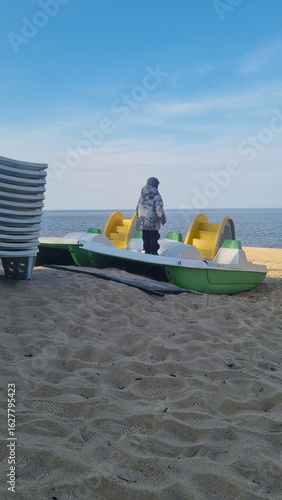 Stack of sun loungers on the beach.  Start or end of the summer season at the resort city. Heap of sunbeds at the coastline. Boy stands on a catamaran.