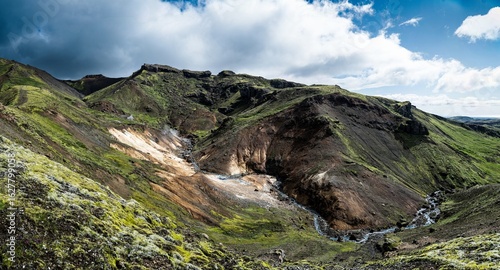 View of rugged, moss-covered hillsides meet barren earth, carved by a winding stream under a dramatic sky, Hveragerdi, GrÃ­msnes- og Grafningshreppur, Iceland.