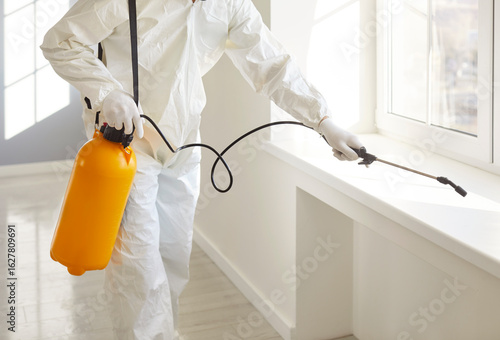 Man exterminator fighting against insects in house spraying poison from sprayer bottle. Worker in protective white ppe suit disinfecting apartments or doing pest control with disinfectant spray