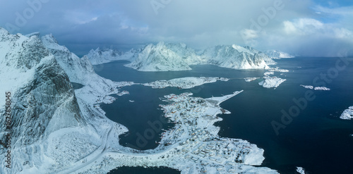 Aerial view of snow-dusted peaks meet the deep blue sea, dotted with islands and coastal villages under a soft, diffused light, Lofoten, Norway.