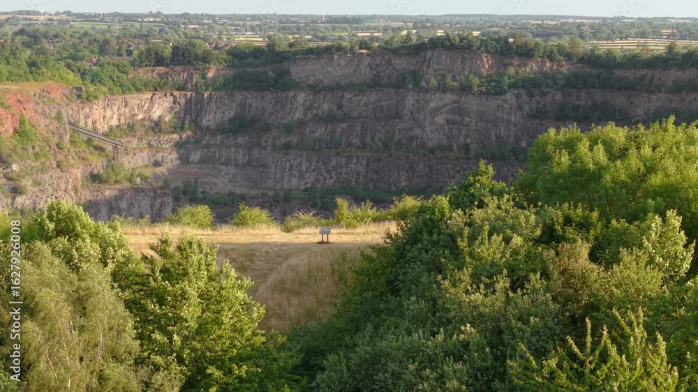 Quarry wall stretching across landscape with terraced rock faces and forest rim. Excavation edge running parallel to greenery with stratified stone formations. Mining site bordering natural growth