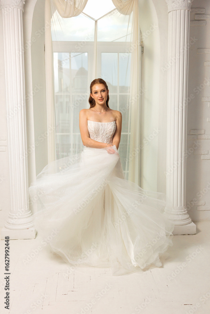Elegant bride in flowing wedding dress poses near window in bridal studio for a timeless portrait Elegant bride in flowing wedding dress poses near window in bridal studio for a timeless portrait