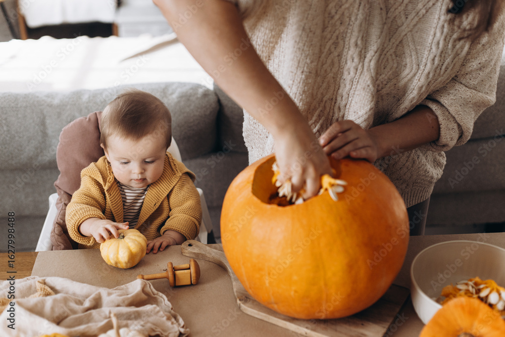 Fototapeta premium Happy Halloween. Happy mother carving halloween pumpkin together with baby son on table with spooky halloween decor. Family making jack o lantern, holiday joyful time