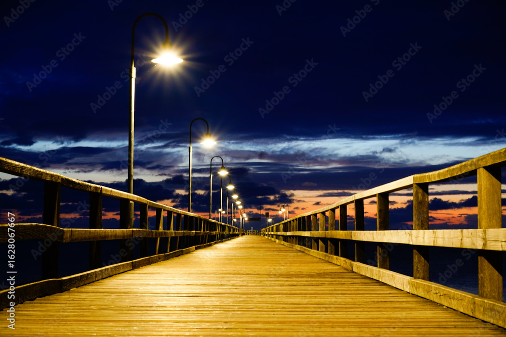 Obraz premium An illuminated pier disappears into the darkness, lanterns illuminating the way under a dramatically cloudy sky. Sun and clouds at dawn on the Baltic Sea beach of 18586 Göhren, Rügen Island