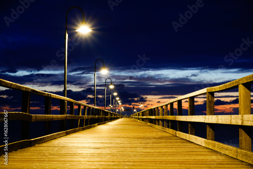 Fototapeta Naklejka Na Ścianę i Meble -  An illuminated pier disappears into the darkness, lanterns illuminating the way under a dramatically cloudy sky. Sun and clouds at dawn on the Baltic Sea beach of 18586 Göhren, Rügen Island