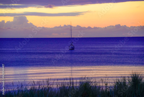 Fototapeta Naklejka Na Ścianę i Meble -  A lone sailboat drifts on the calm Baltic Sea at sunrise, surrounded by purple and orange clouds. Sun and clouds at dawn on the Baltic Sea beach of 18586 Göhren, Rügen Island