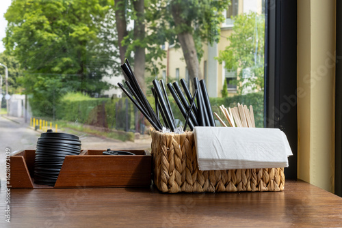 Fotografie Set of black plastic straws, wooden stirrers, napkins and cup lids in containers on wooden counter near window