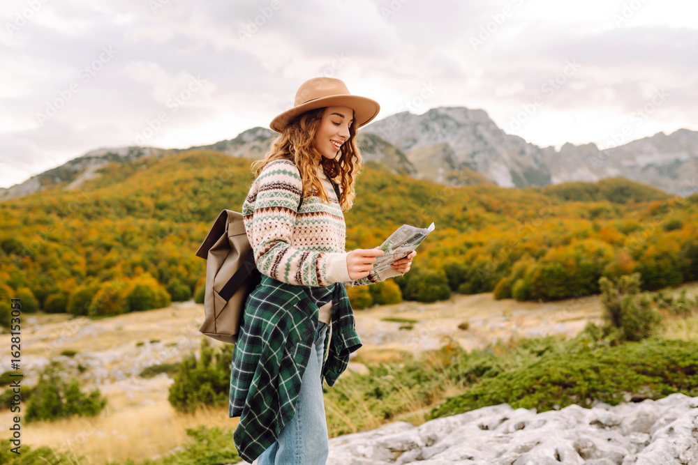 Fototapeta premium Smiling woman in backpack holding map against majestic mountains background. Young female traveler enjoying landscapes with map of area and choosing direction for hiking. Travel concept, navigation.