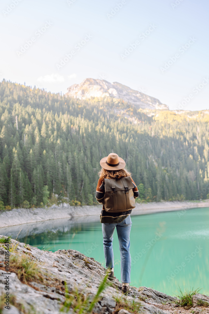 Naklejka premium A back view of a female hiker in a hat and with a backpack sitting on a rock overlooking a high mountain lake. Young woman feels freedom and enjoys mountain landscape. Hiking and nature concept.