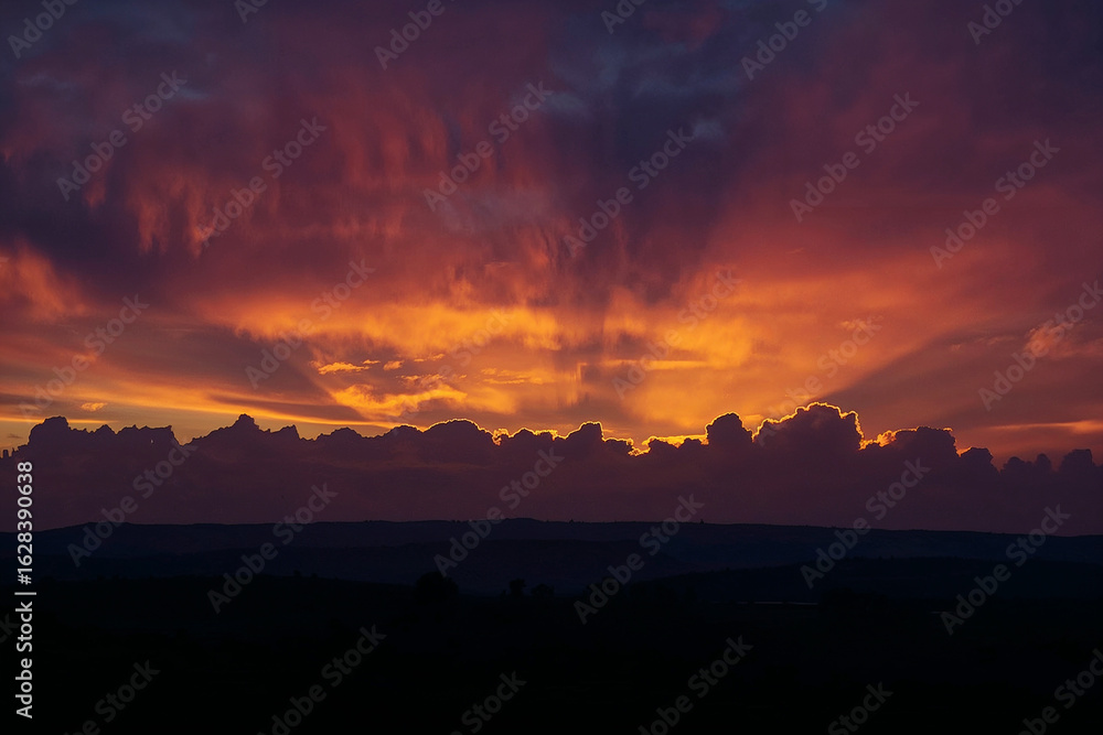 Fototapeta premium Fiery Sunset with Sun Rays Breaking Through Clouds Over Silhouetted Landscape dramatic