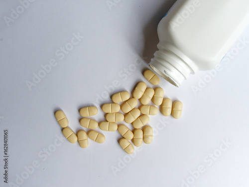 Yellow tablets spilled from a white bottle onto a plain white surface. Medical, pharmaceutical, and health concepts.