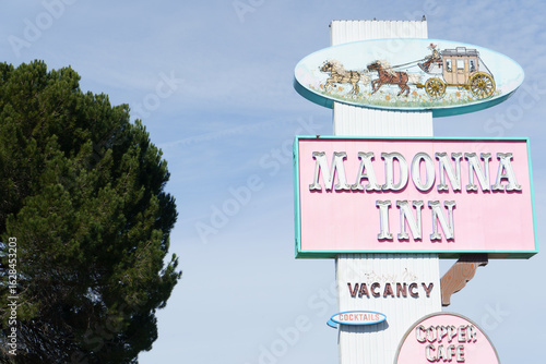 Carta da parati The iconic pink roadside sign for the Madonna Inn, featuring a vintage horse-drawn carriage illustration, stands tall against a bright blue sky
