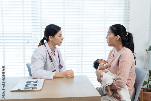 Mother holding her newborn baby while consulting with a female pediatrician in a bright medical office, discussing child health care and postpartum wellness for newborn development and family health.