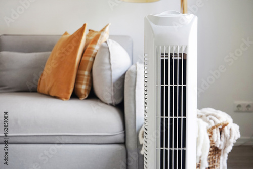 White tower fan against the background of a sofa and a basket with blankets in a modern light interior