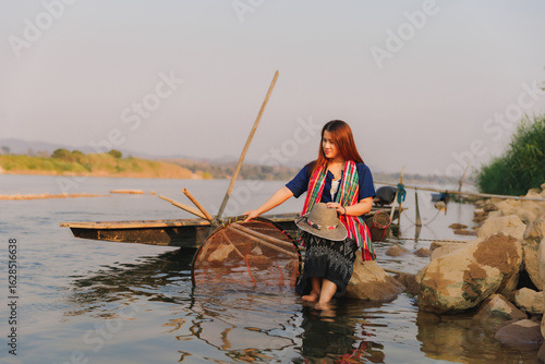 Fototapeta A woman sits in the shallows of the river with her fishing equipment