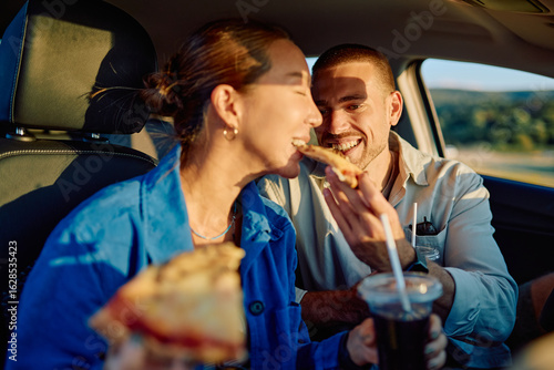 Happy couple enjoying pizza and drinks while sharing laughter and love during a memorable road trip inside their car at sunset