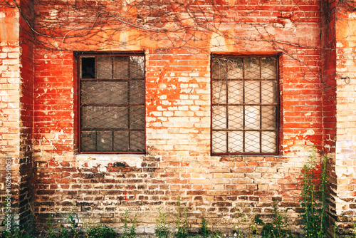 Fotografi Old brick wall with rusty barred windows, cracked plaster, and creeping dry vine