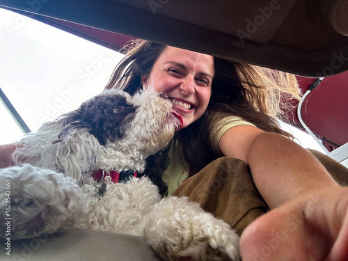 A low-angle selfie captures a joyful woman smiling as her fluffy white dog affectionately licks her face, showing their close bond.